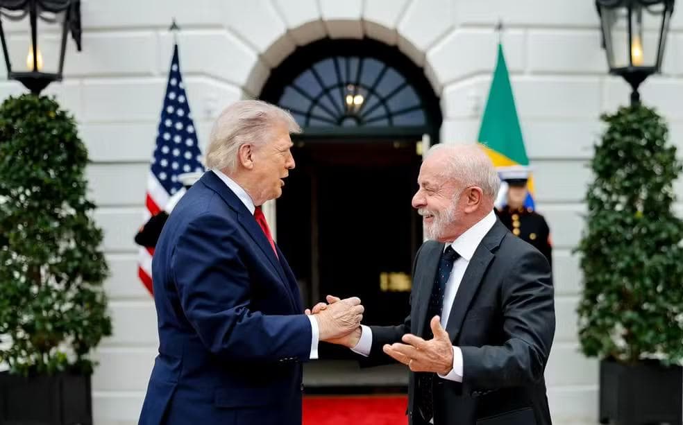 An outdoor photograph at the entrance of the White House (North Portico). Donald Trump (L) and Lula da Silva (R) are shaking hands and smiling, standing on a red carpet. US and Brazilian flags are visible behind them.