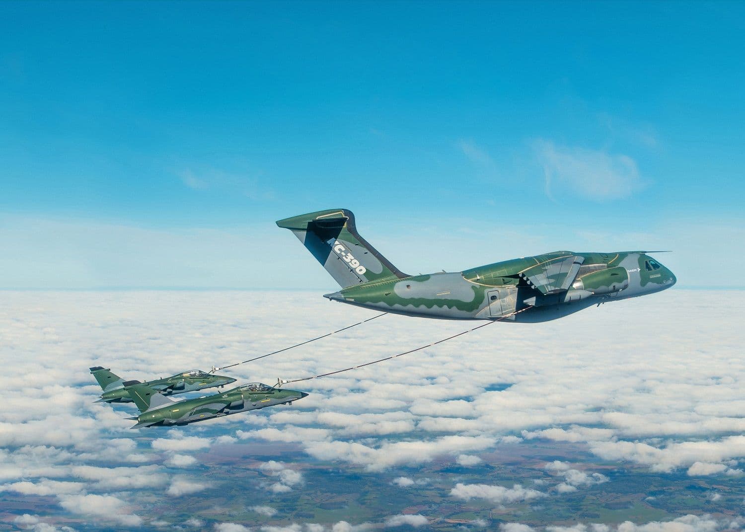 An Embraer KC-390 Millennium military transport aircraft in green camouflage conducting a mid-air refueling operation with two fighter jets above the clouds.