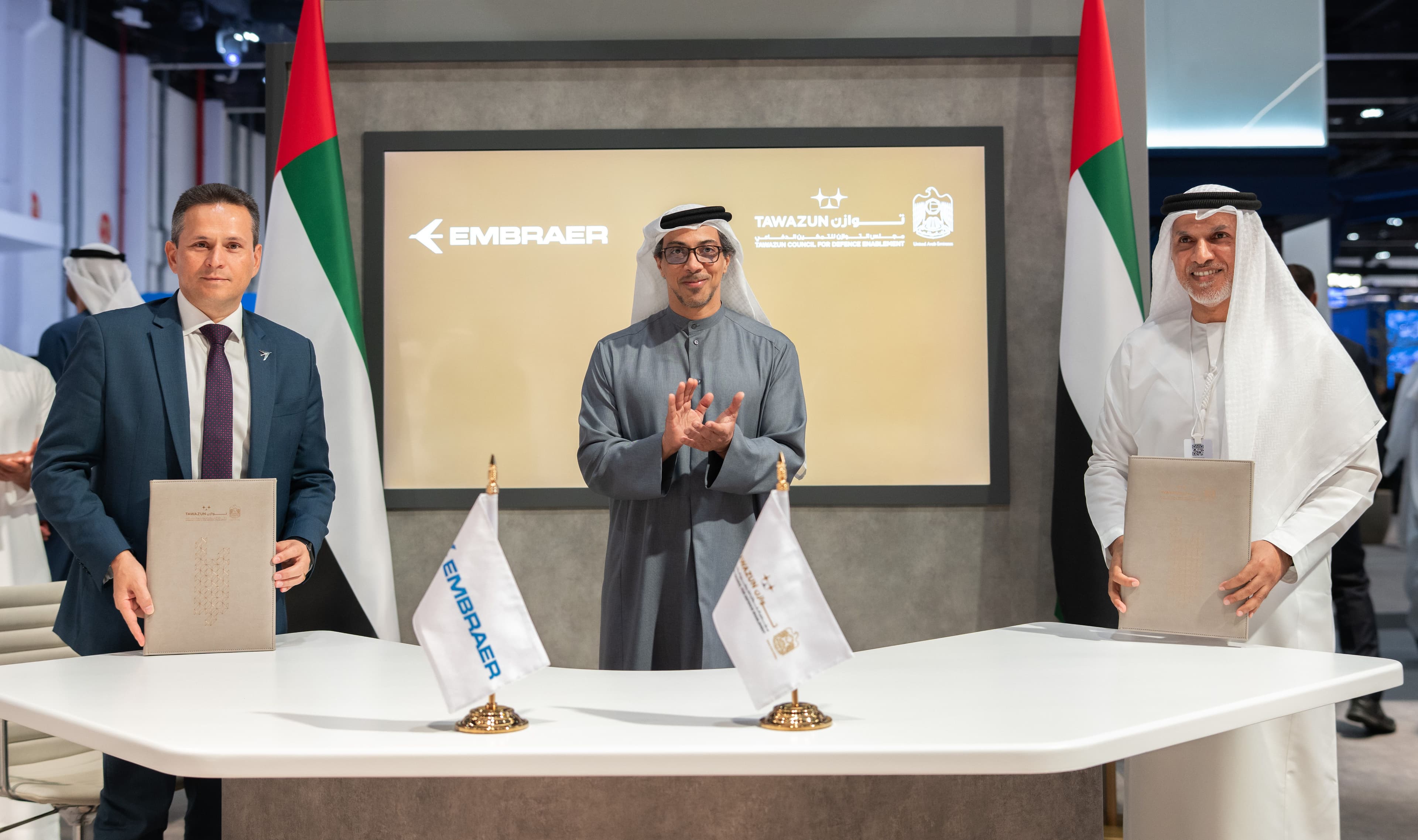 Bosco da Costa Junior of Embraer (left) and H.E. Dr. Nasser Humaid Al Nuaimi of the Tawazun Council (right) holding signed official documents. In the center, H.H. Sheikh Mansour bin Zayed Al Nahyan claps in celebration. They are standing behind a white table with Embraer and Tawazun flags, with UAE flags and corporate logos in the background.