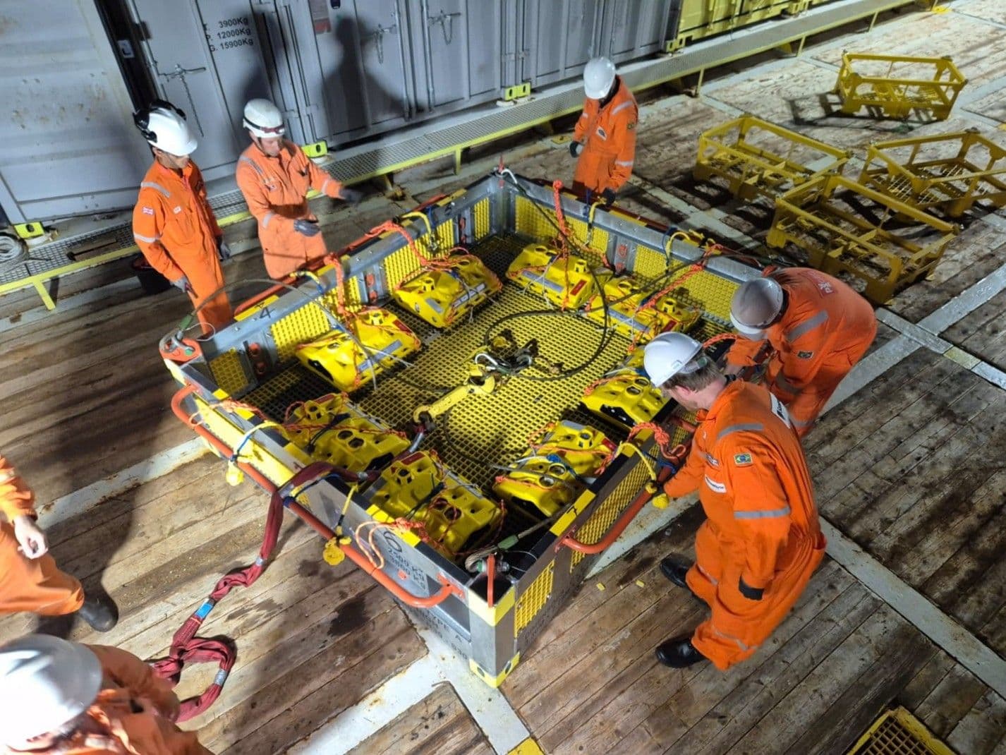 An overhead view of Petrobras and Shell technical crew in orange coveralls and hard hats working on an offshore vessel deck. They are inspecting and securing a fleet of yellow OD OBN (On-demand Ocean Bottom Nodes) subsea seismic sensors in a large metal deployment cage, destined for deployment in Brazil's pre-salt Mero field.