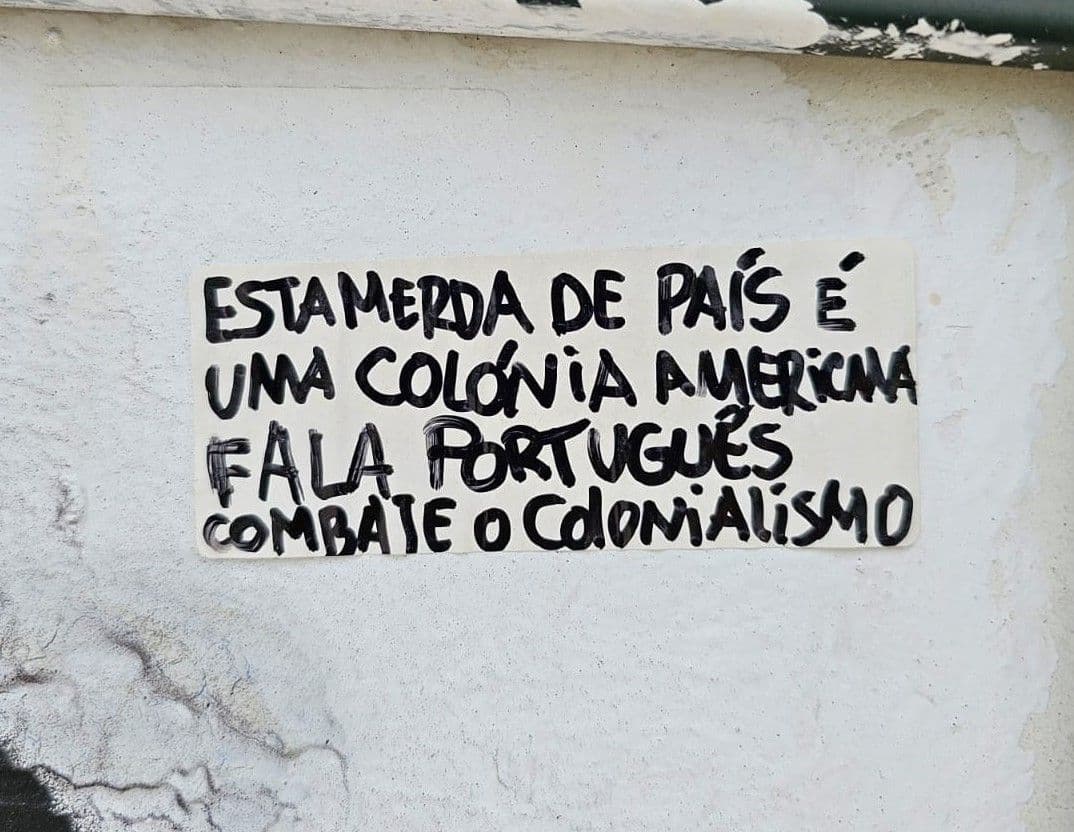 Close-up photo of a handwritten sticker pasted on a white wall in Lisbon. The text is in Portuguese and reads: “Esta merda de país é uma colónia americana. Fala português. Combate o colonialismo,” which translates to: “This shitty country is an American colony. Speak Portuguese. Fight colonialism.” The image was taken near a tourist attraction.