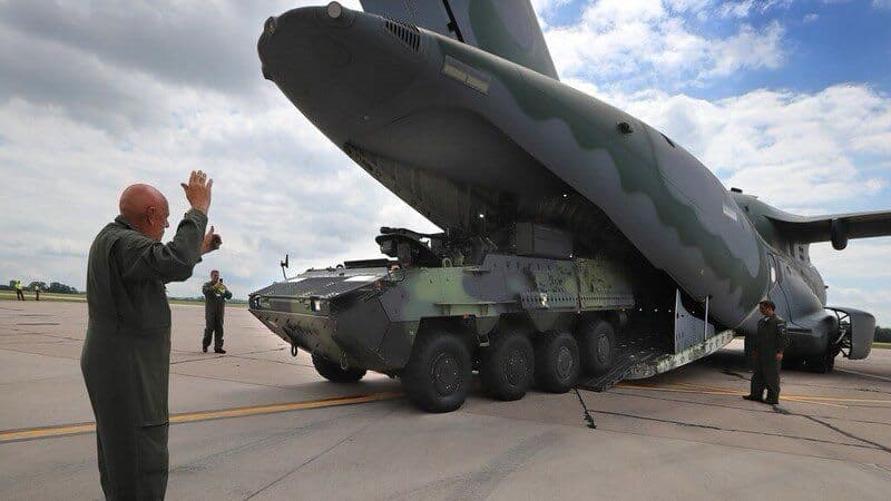 A heavy 8x8 military armored vehicle driving up the rear cargo ramp to be loaded into the spacious cargo bay of an Embraer C-390 Millennium, guided by a crew member using hand signals.